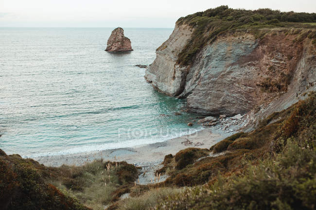 Tranquillo paesaggio panoramico con scogliera rocciosa coperta di erba verde e mare calmo con roccia solitaria tra l'acqua nella giornata nuvolosa — Foto stock