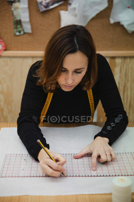 From above focused adult woman sitting at table and making cutout while working in professional dressmaking studio — Stock Photo
