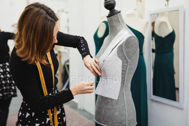 Woman with tape measure attaching paper cutout to mannequin with pin while making clothes in professional workshop — Stock Photo