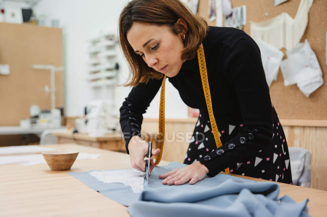 Mujer que usa tijeras afiladas para cortar el detalle de la prenda de tela mientras está sentada en la mesa en el taller de sastrería - foto de stock