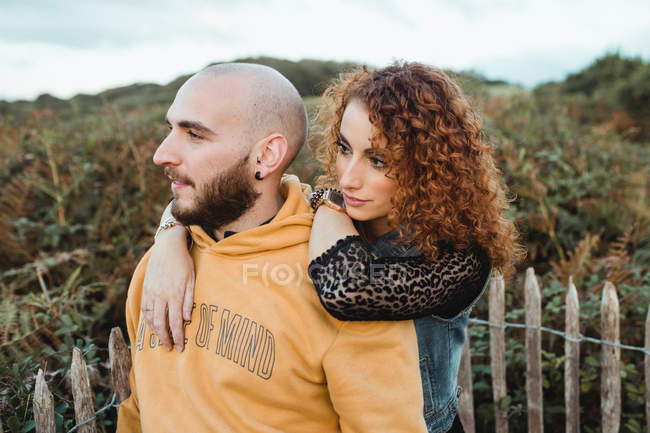 Jovem de capuz amarelo abraçando a namorada em vestido e colete de ganga enquanto estava perto de cerca juntos no prado — Fotografia de Stock