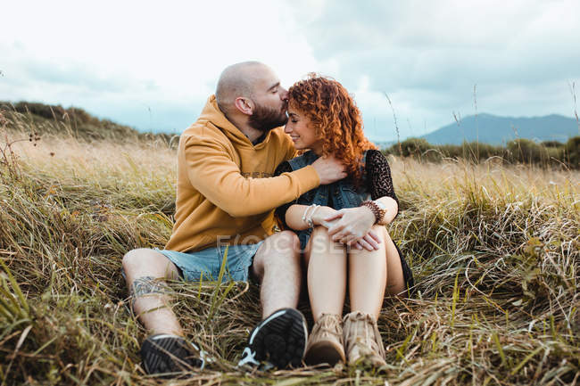 Jeune homme en sweat à capuche jaune embrassant petite amie en robe et gilet en denim tout en se tenant près de la clôture ensemble sur la prairie, baisers — Photo de stock