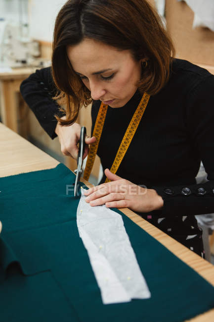Donna che usa forbici affilate per tagliare il dettaglio dell'indumento dal tessuto mentre siede a tavola in laboratorio sartoriale — Foto stock