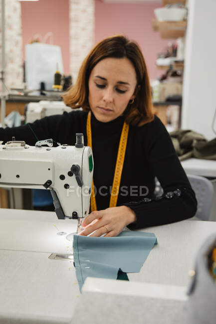 Mulher adulta sentada à mesa e fazendo peça de vestuário na máquina de costura enquanto trabalhava em estúdio profissional — Fotografia de Stock