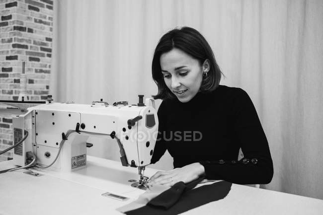 Доросла жінка сидить за столом і робить частину одягу на швейній машині під час роботи в професійній студії — стокове фото