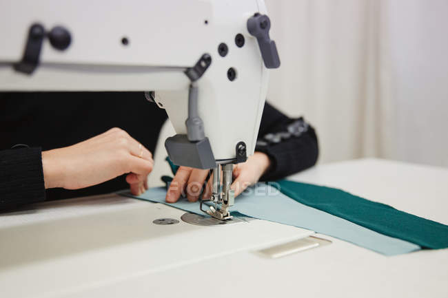 Mujer sentada en la mesa y haciendo parte de la prenda en la máquina de coser mientras trabaja en un estudio profesional - foto de stock