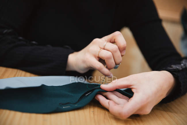 Primer plano de modista usando aguja e hilo para coser ropa personalizada sobre la mesa en taller profesional - foto de stock