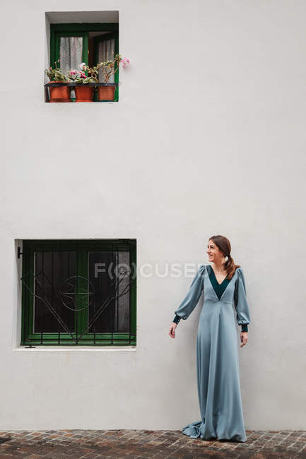 Fêmea feliz em vestido vintage sorrindo e olhando para longe enquanto estava em pé no pavimento fora da casa velha — Fotografia de Stock