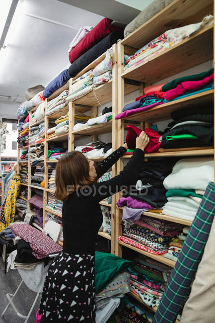 Vista lateral da mulher adulta vasculhando a prateleira ao selecionar pedaço de pano para o trabalho na sala de armazenamento da oficina de alfaiate — Fotografia de Stock