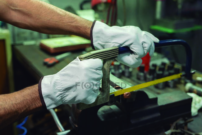 Mains gantées d'un réparateur professionnel anonyme sciant des détails métalliques avec une scie à métaux pendant le travail en usine — Photo de stock