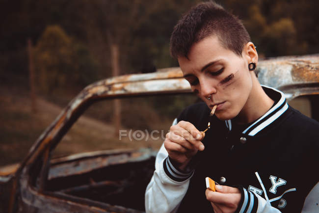 Jeune femme aux cheveux courts et peint cigarette éclairage visage avec briquet tout en se tenant près de vieille voiture rouillée dans la campagne — Photo de stock