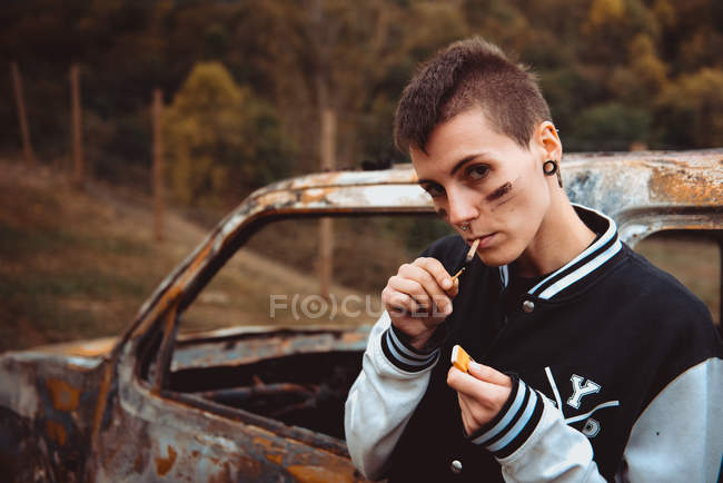Молодая женщина с короткими волосами и раскрашенным лицом, зажигая сигарету с зажиганием, стоя рядом со старой ржавой машиной в сельской местности — стоковое фото