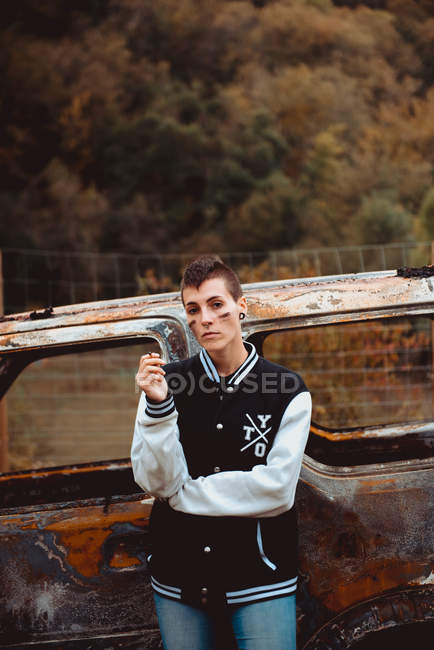 Young woman with short hair smoking cigarette while resting near aged burnt vehicle in countryside, looking in camera — Stock Photo