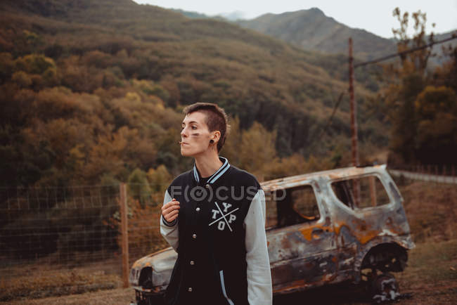 Rebel woman with short hair smoking cigarette near old burnt auto in countryside — Stock Photo