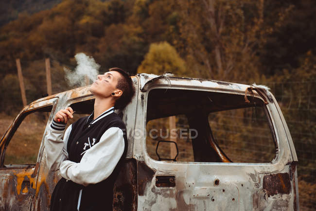 Стильний бойфренд з коротким волоссям і пофарбованою сигаретою для куріння обличчя як спираючись на старий іржавий автомобіль у сільській місцевості — стокове фото