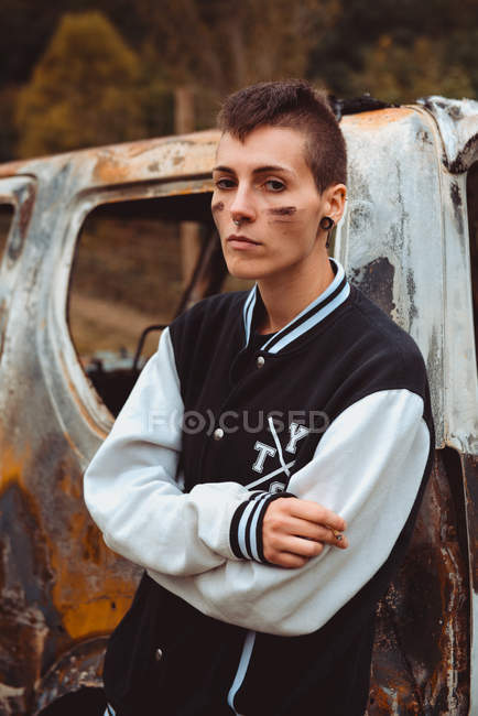Elegante maschiaccio con i capelli corti e volto dipinto fumare sigaretta come appoggiato su vecchia auto arrugginita in campagna — Foto stock