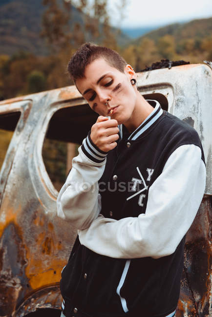 Stylish tomboy with short hair and painted face smoking cigarette as leaning on old rusty car in countryside — Stock Photo