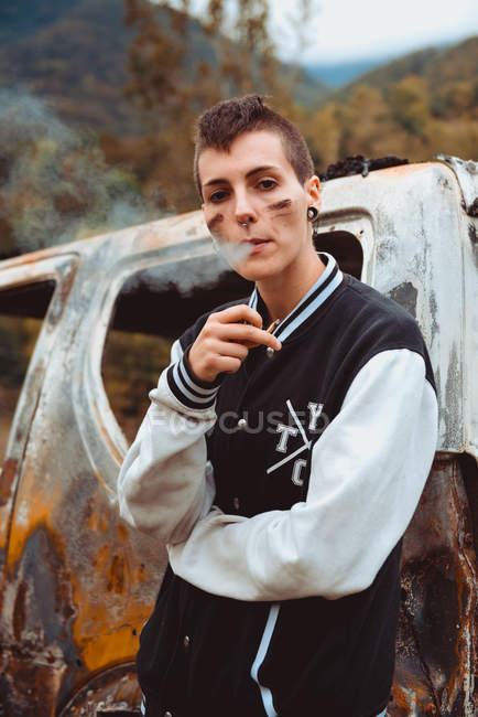 Молода жінка з коротким волоссям курить сигарету, відпочиваючи біля старого спаленого автомобіля в сільській місцевості, дивлячись в камеру — стокове фото