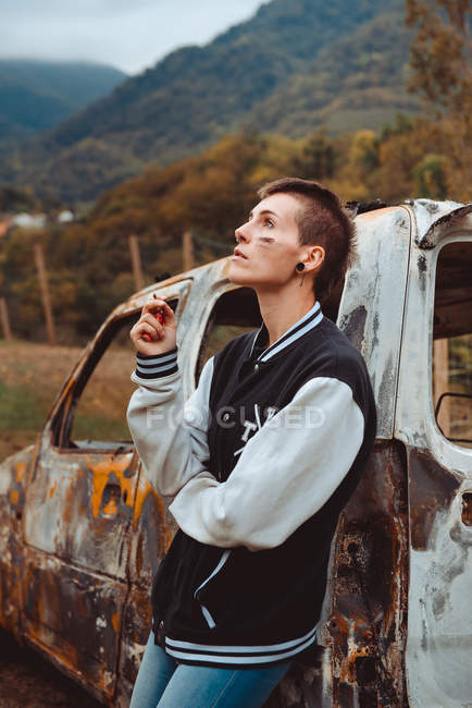 Jovem fêmea com cabelo curto fumando cigarro enquanto descansa perto de veículo queimado envelhecido no campo — Fotografia de Stock