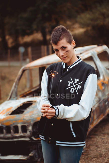 Young woman with short hair holding burning sparkler and looking in camera while standing near old rusty vehicle in countryside — Stock Photo