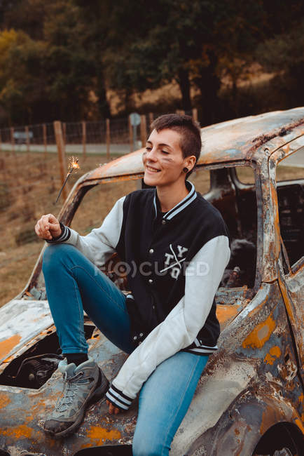 Jovem mulher com cabelo curto segurando queimando sparkler e olhando para longe enquanto sentado no velho veículo enferrujado no campo — Fotografia de Stock