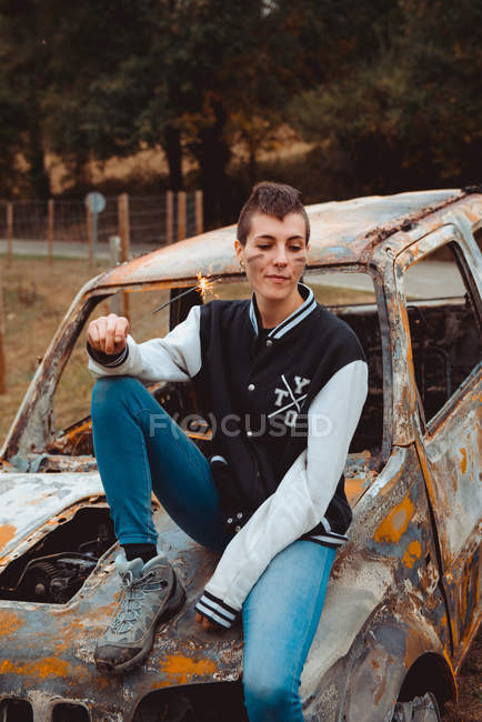 Jeune femme aux cheveux courts tenant étincelant brûlant et regardant loin tout en étant assis sur un vieux véhicule rouillé dans la campagne — Photo de stock