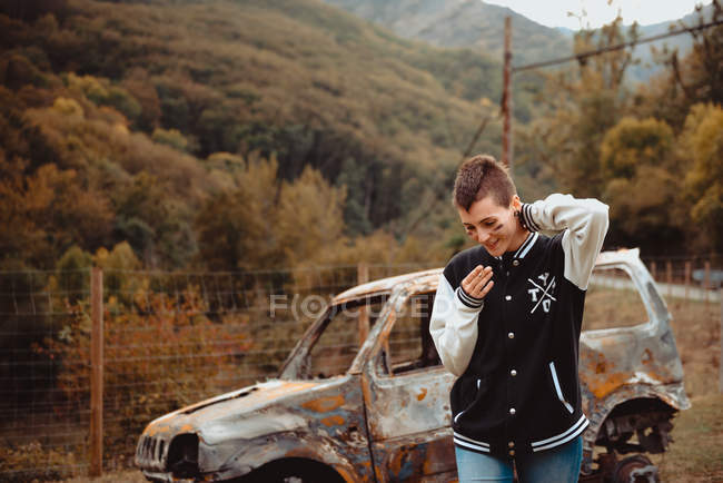 Jovem fêmea com cabelo curto fumando cigarro enquanto caminhava perto de veículo queimado envelhecido no campo — Fotografia de Stock