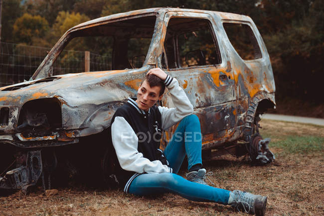 Jovem fêmea em roupa casual tocando cabelo curto e olhando para longe, sentado no chão perto de carro velho queimado no campo — Fotografia de Stock