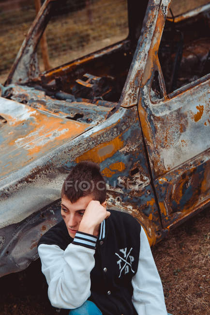 Молодая женщина в повседневной одежде трогает короткие волосы и смотрит в сторону, сидя на земле рядом со старым сожженным автомобилем в сельской местности — стоковое фото