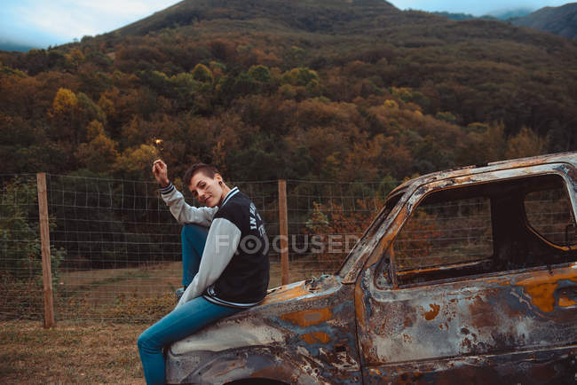 Junge Frau mit kurzen Haaren hält brennende Wunderkerze in der Hand und blickt in die Kamera, während sie auf einem alten rostigen Fahrzeug auf dem Land sitzt — Stockfoto