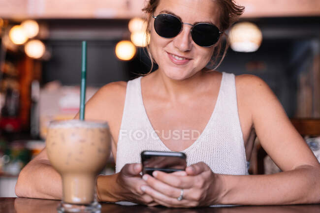 Mujer alegre en gafas de sol navegando teléfono inteligente en la cafetería - foto de stock