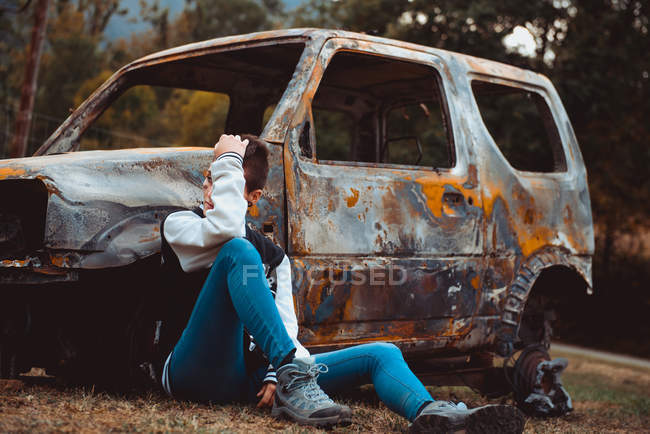 Молодая женщина в повседневной одежде трогает короткие волосы и смотрит в сторону, сидя на земле рядом со старым сожженным автомобилем в сельской местности — стоковое фото