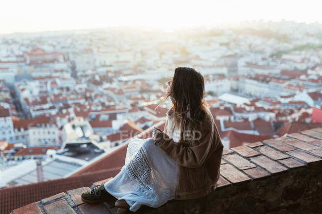 Vue latérale de la jeune femme en veste et robe assise sur une barrière de briques et admirant le paysage urbain au matin ensoleillé au Portugal — Photo de stock