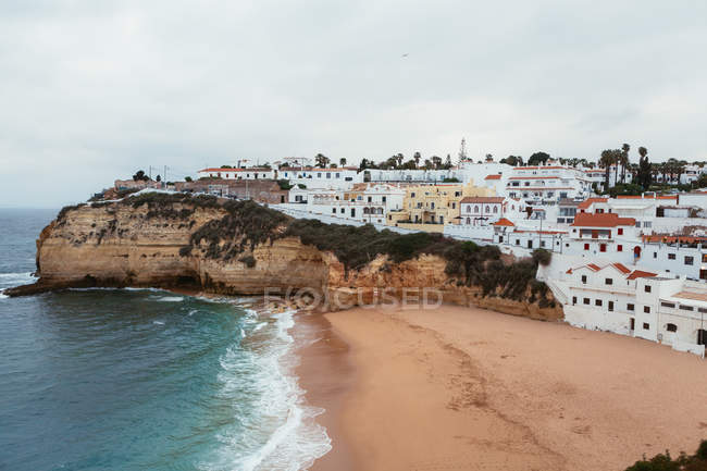 Mare tempestoso che ondeggia vicino a ruvida scogliera e spiaggia sabbiosa con piccola città costiera nella giornata nuvolosa in Portogallo — Foto stock