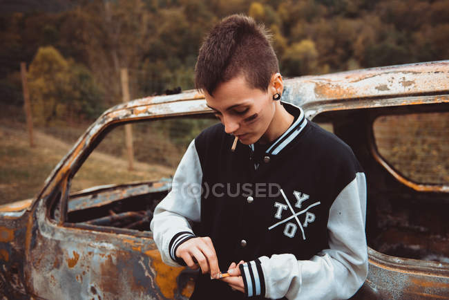 Young woman with short hair and painted face lighting cigarette standing near old rusty car in countryside — Stock Photo