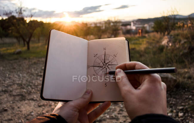 Unrecognizable person drawing sketch of compass in notebook during trip through countryside in evening — Stock Photo