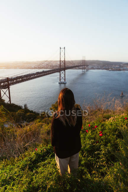 Visão traseira da mulher em pé na grama e olhando para ponte suspensa sobre o rio na noite sem nuvens em Portugal — Fotografia de Stock
