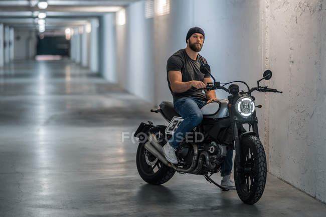 Plein corps barbu gars en vêtements décontractés assis sur la moto et regardant la caméra dans le couloir du garage moderne — Photo de stock