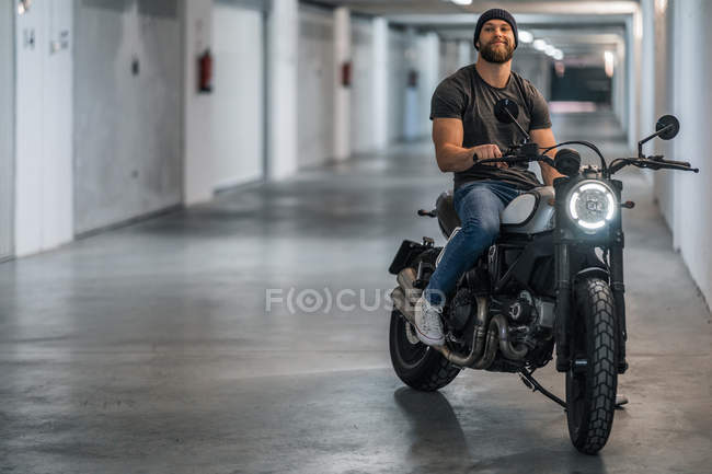 Full body bearded guy in casual clothes sitting on motorcycle and looking at camera in corridor of modern garage — Stock Photo