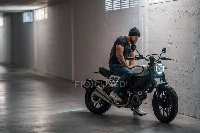 Plein corps barbu gars en vêtements décontractés assis sur la moto et regardant loin dans le couloir du garage moderne — Photo de stock