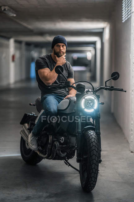 Tipo barbudo de cuerpo completo en ropa casual sentado en motocicleta y mirando a la cámara en el pasillo de garaje moderno - foto de stock