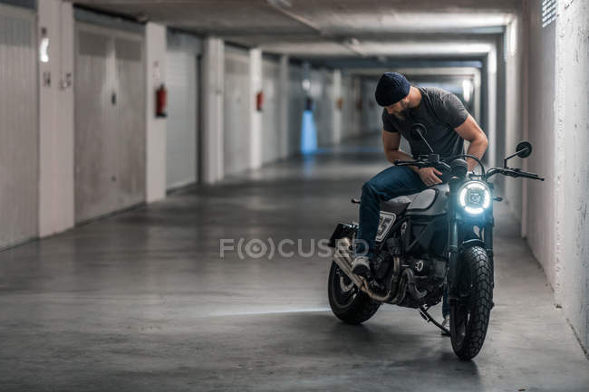 Pieno corpo barbuto ragazzo in abiti casual seduto sulla moto e guardando lontano nel corridoio del garage moderno — Foto stock