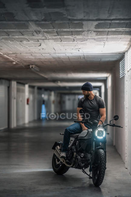 Tipo barbudo de cuerpo completo en ropa casual sentado en motocicleta y mirando hacia otro lado en el pasillo de garaje moderno - foto de stock