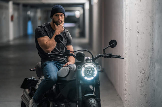 Corpo inteiro barbudo cara em roupas casuais sentado na motocicleta e olhando para a câmera no corredor da garagem moderna — Fotografia de Stock