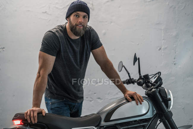 Corpo inteiro barbudo cara em roupas casuais de pé perto de moto e olhando para a câmera no corredor da garagem moderna — Fotografia de Stock