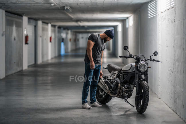 Corpo inteiro barbudo cara em roupas casuais de pé perto de moto no corredor da garagem moderna — Fotografia de Stock