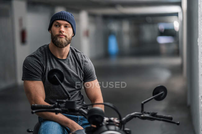 Pieno corpo barbuto ragazzo in abiti casual seduto sulla moto e guardando la fotocamera nel corridoio del garage moderno — Foto stock