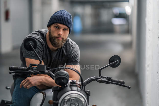 Tipo barbudo de cuerpo completo en ropa casual sentado en motocicleta y mirando a la cámara en el pasillo de garaje moderno - foto de stock