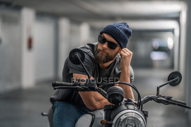 Corpo inteiro barbudo cara em roupas casuais e óculos de sol na motocicleta no corredor da garagem moderna — Fotografia de Stock