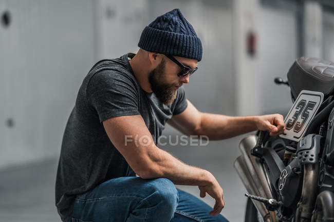 Hombre barbudo en sombrero levantando la ceja y mirando a la cámara mientras está sentado cerca de la motocicleta en el fondo borroso del garaje contemporáneo - foto de stock